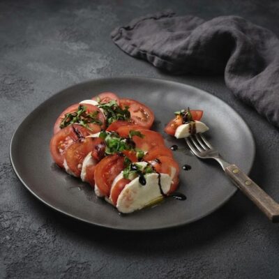 Caprese salát z čerstvých rajčat, mozzarelly a bazalky s olivovým olejem a kapkami tmavého balzamikového krému na šedém talíři.